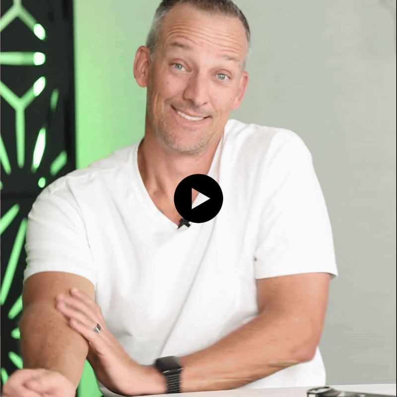 Man with short gray hair and a beard, wearing a white t-shirt, sitting at a table, smiling, with a play button over the image.